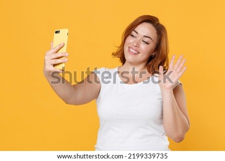 Smiling young redhead plus size body positive female woman girl in white casual t-shirt doing selfie shot on mobile phone waving greeting with hand isolated on yellow color background studio portrait