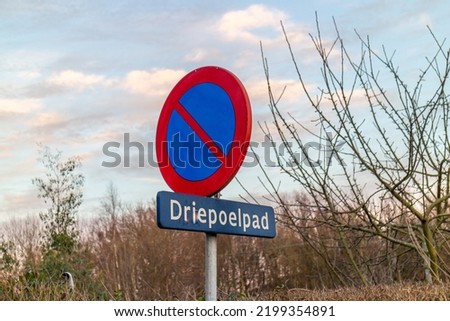 Traffic signs: Parking prohibited, street name Driepoelpad against cloudy sky, round plate with red and blue colors, bare tree branches background, Sittard-Geleen, South Limburg, the Netherlands