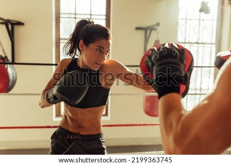 Pretty young woman trains in boxing ring with partner near red punching bag and other sparring equipment Royalty-Free Stock Photo #2199363745