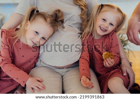 Cropped shoot of two little blonde girls with tails sitting with their pregnant mom, happy family waiting for a newborn. Girls at the kitchen with pregnant mom. Kids, health care and pregnancy.