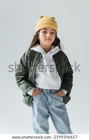 portrait of cute kid in yellow beanie hat and stylish autumnal outfit posing with hands in pockets isolated on grey Royalty-Free Stock Photo #2199396657