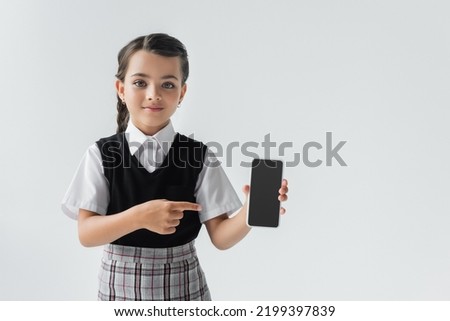 cute schoolgirl in uniform pointing with finger at smartphone with blank screen isolated on grey