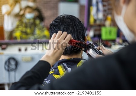 A close-up view of a man's back is a masked barber woman using a comb and clippers to professionally style a man's hair inside a barbershop room with mirrors and a classic pattern. Royalty-Free Stock Photo #2199401155