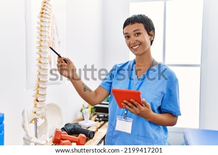 Young hispanic woman wearing physiotherapist uniform pointing to vertebral column using touchpad at physiotherapy clinic Royalty-Free Stock Photo #2199417201