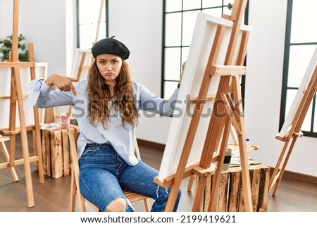 Young hispanic artist woman painting on canvas at art studio pointing down looking sad and upset, indicating direction with fingers, unhappy and depressed. 