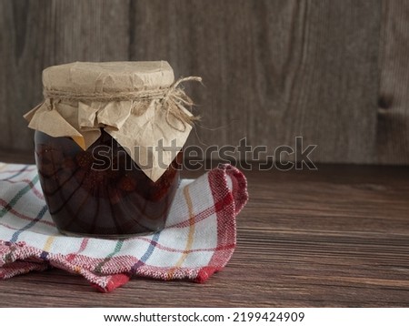 A jar of homemade strawberry jam covered with parchment on a wooden background with a kitchen towel with a place for text. Winter preparations, sweets for tea.