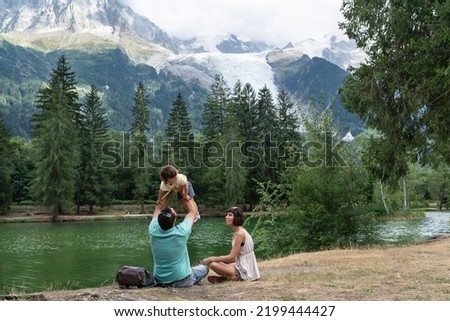 Father holding her little daughter up while mother is looking at them, both sitting on a shore lake with the majestic Mont blanc on the background.