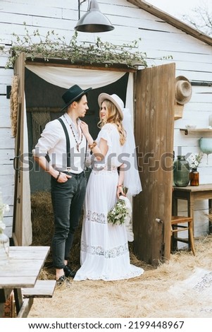young couple newlyweds bride in short white dress and wreath with veil and groom in hat and jeans with suspenders hugging and having fun at barn of country house 