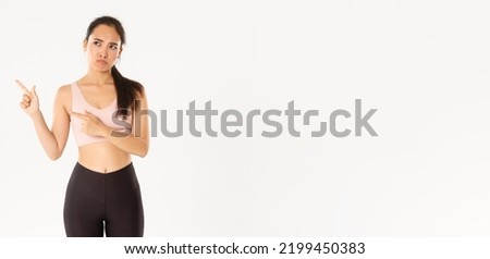 Sport, wellbeing and active lifestyle concept. Complaining sad asian girl in activewear, sulking and frowning upset as pointing upper left corner, end of sale offer, standing white background