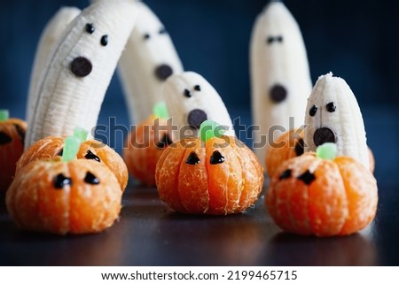 Halloween cute pumpkin orange fruit with banana ghosts with chocolate faces behind them. Healthy snack with funny face for child's party decoration. Selective focus on center with blurred background.