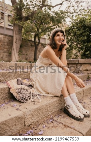 Cute young caucasian girl posing sitting on steps relaxing outdoors. Brunette wears sundress in summer. Leisure concept