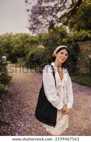 Beautiful young caucasian brunette girl looks at camera, walks through flowering garden. Model wears headscarf, bag and dress in summer. Rest time concept