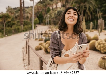 Nice young caucasian girl browsing social media on smartphone relaxing outdoors. Brunette wears summer dress in warm weather. Addiction concept