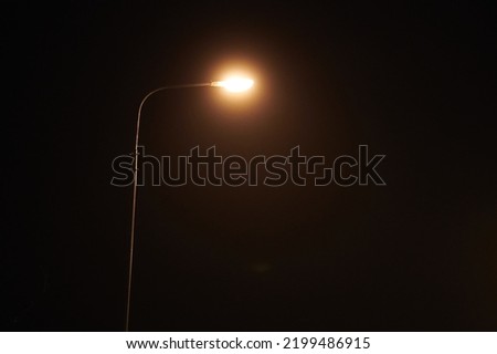 One night lamppost shines with faint mysterious yellow light through evening fog. Streetlight shine at quiet city night, magic atmospheric light in mystical darkness, copy space Royalty-Free Stock Photo #2199486915