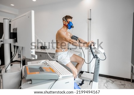 Man athlete with breath mask and electrodes training on bike simulator, examining his cardiovascular system at medical office Royalty-Free Stock Photo #2199494167