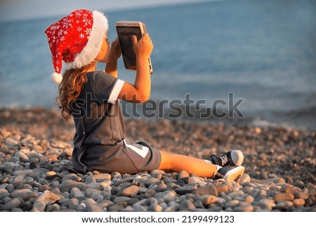 a little girl in a blue dress and a red santa claus hat sits on the seashore in the sunset and watches cartoons on a tablet, plays and chats online with friends