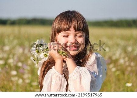 Portrait of a little smiling girl in a white dress on a chamomile field on a bright sunny summer day