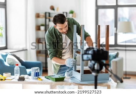 blogging, furniture restoration and home improvement concept - man or blogger with camera repainting old wooden table with paint roller in grey color and recording tutorial video Royalty-Free Stock Photo #2199527523