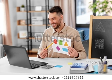 distance education, primary school and remote job concept - happy smiling male teacher with laptop and picture of geometric shapes in different colors having online class at home office