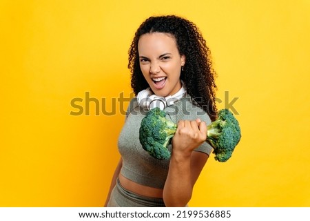 Happy excited active funny hispanic or brazilian curly woman in sport outfit, holds exercise with broccoli dumbbell while standing on isolated orange background, smiling, emotional facial expression Royalty-Free Stock Photo #2199536885