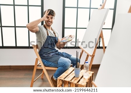 Young hispanic man painting at art studio smiling happy doing ok sign with hand on eye looking through fingers 