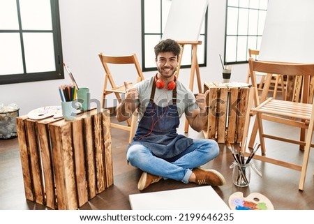 Young hispanic man sitting at art studio success sign doing positive gesture with hand, thumbs up smiling and happy. cheerful expression and winner gesture. 
