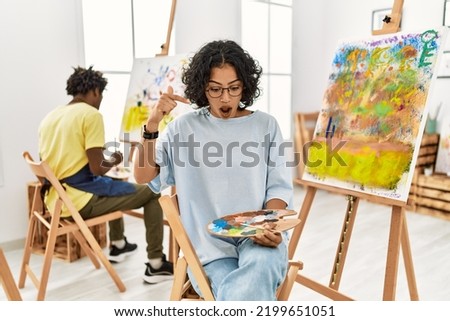 Young hispanic woman at art studio pointing down with fingers showing advertisement, surprised face and open mouth 