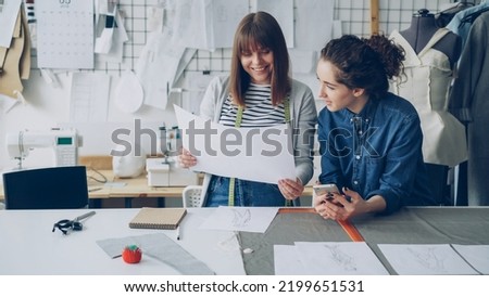 Attractive young women working in design industry are comparing sketches and talking about them while working in modern tailor shop. Productive teamwork concept.