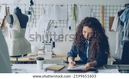 Young woman fashion designer is drawing ladies' garment sketch at desk in modern workshop. Mannequin, sewing items, to-go coffee are visible. Creativity in clothing manufacturing concept.