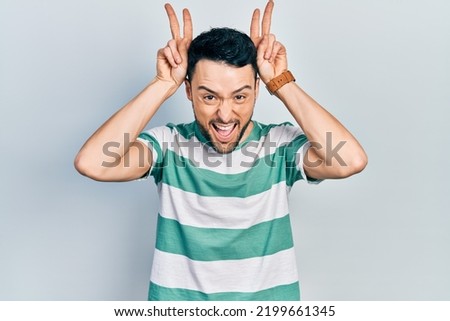 Young hispanic man wearing casual clothes posing funny and crazy with fingers on head as bunny ears, smiling cheerful 