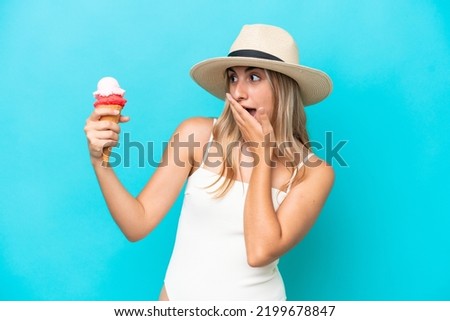 Young caucasian woman in swimsuit with a cornet ice cream isolated on blue background with surprise and shocked facial expression