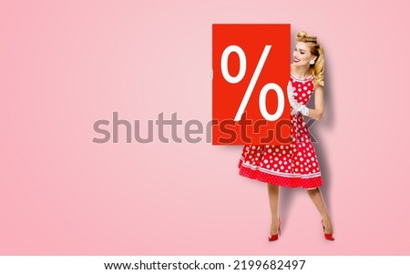 Beautiful woman in pinup dress holding, showing red board with procents % sign. Full body pin up girl with advertising offer isolated on rose pink background. Sales, discounts rebates deals ad concept