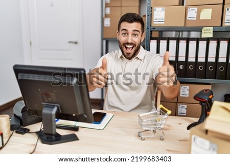 Handsome hispanic man working at small business commerce approving doing positive gesture with hand, thumbs up smiling and happy for success. winner gesture. 