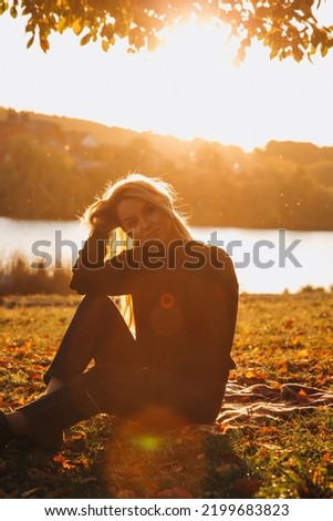 Beautiful girl with long blond hair in autumn landscape at sunset. Autumn portrait of a girl sitting on the grass, front view, selective focus
