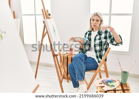 Young artist woman painting on canvas at art studio pointing down looking sad and upset, indicating direction with fingers, unhappy and depressed. 