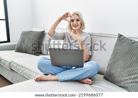 Young caucasian woman using laptop at home sitting on the sofa smiling making frame with hands and fingers with happy face. creativity and photography concept. 