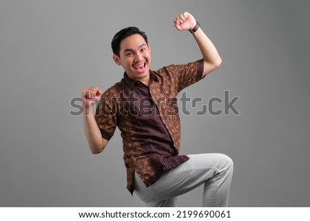 Excited young Asian man wearing batik shirt celebrating victory and raised fists isolated on grey background Royalty-Free Stock Photo #2199690061