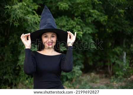 Halloween. A young beautiful woman in a witch's hat and a black dress in the park. Space for text.