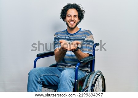 Handsome hispanic man sitting on wheelchair smiling with hands palms together receiving or giving gesture. hold and protection 