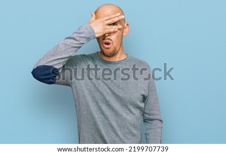 Bald man with beard wearing casual clothes peeking in shock covering face and eyes with hand, looking through fingers with embarrassed expression. 