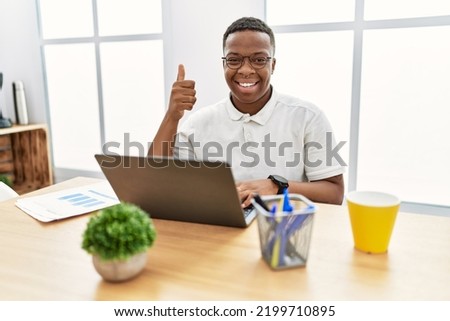 Young african man working at the office using computer laptop doing happy thumbs up gesture with hand. approving expression looking at the camera showing success. 