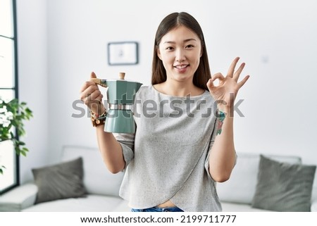 Young asian woman holding italian coffee maker doing ok sign with fingers, smiling friendly gesturing excellent symbol 