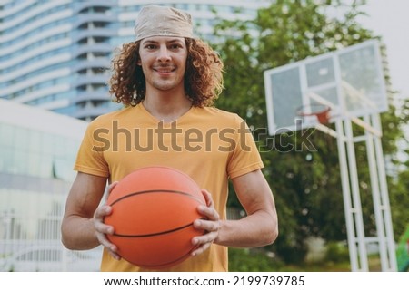 Young happy fun cool man 20s wearing yellow t-shirt bandana look camera holding ball playing basketball on playground in free time rest in city outdoors on open air. Urban lifestyle leisure concept.