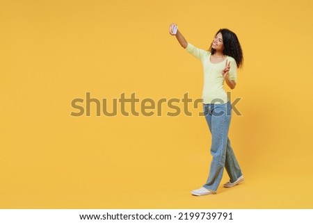 Full size body length overjoyed young happy brunette blogger woman 20s she wears green shirt go walk do selfie shot on mobile cell phone show victory sign isolated on yellow background studio portrait
