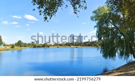 On the shore of the lake grows grass and trees, laid a pedestrian walkway and lampposts installed. The shores are concreted. On the far shore there is an orthodox church and residential houses. Sunny Royalty-Free Stock Photo #2199756279
