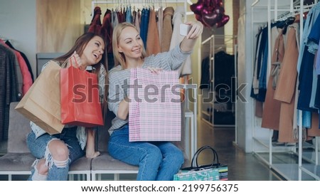 Cute girls are sitting and making selfie with colourful paper bags using smartphone in luxurious women's clothing boutique, then watching pictures together. They are smiling and laughing carelessly.