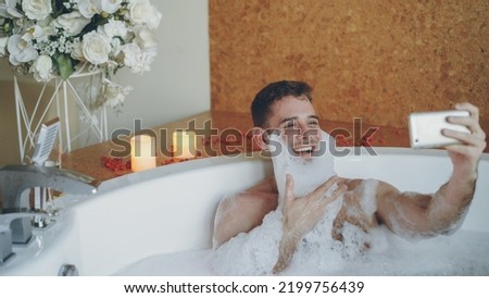 Cheerful young man with foam on beard is taking selfie using smartphone in hot tub in modern spa salon. He is laughing, gesturing, posing and looking at camera.
