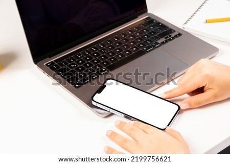 Modern smartphone mock up, selective focus on woman hands holding modern smartphone mock up.  Office desktop set up with laptop and mobile phone. Looking to white blank screen. 