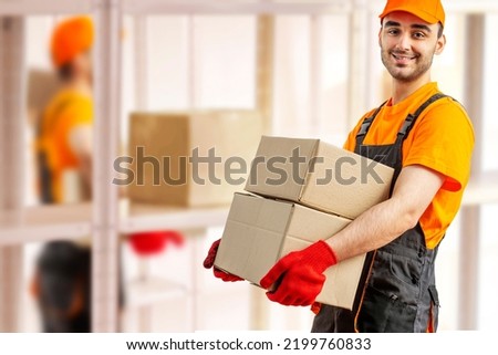 Young man holding cardboard package working in warehouse among racks and shelves. Delivery man with box. Staff laborer, orange uniform cap, t-shirt, coveralls service moving delivering orders goods. 