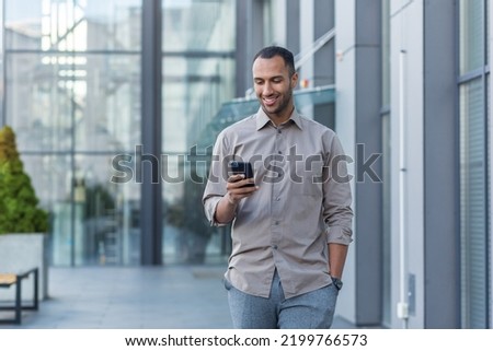 Successful young african american man walking outside office building, engineer software developer programmer smiling and happy using test app on phone, happy satisfied with result. Royalty-Free Stock Photo #2199766573